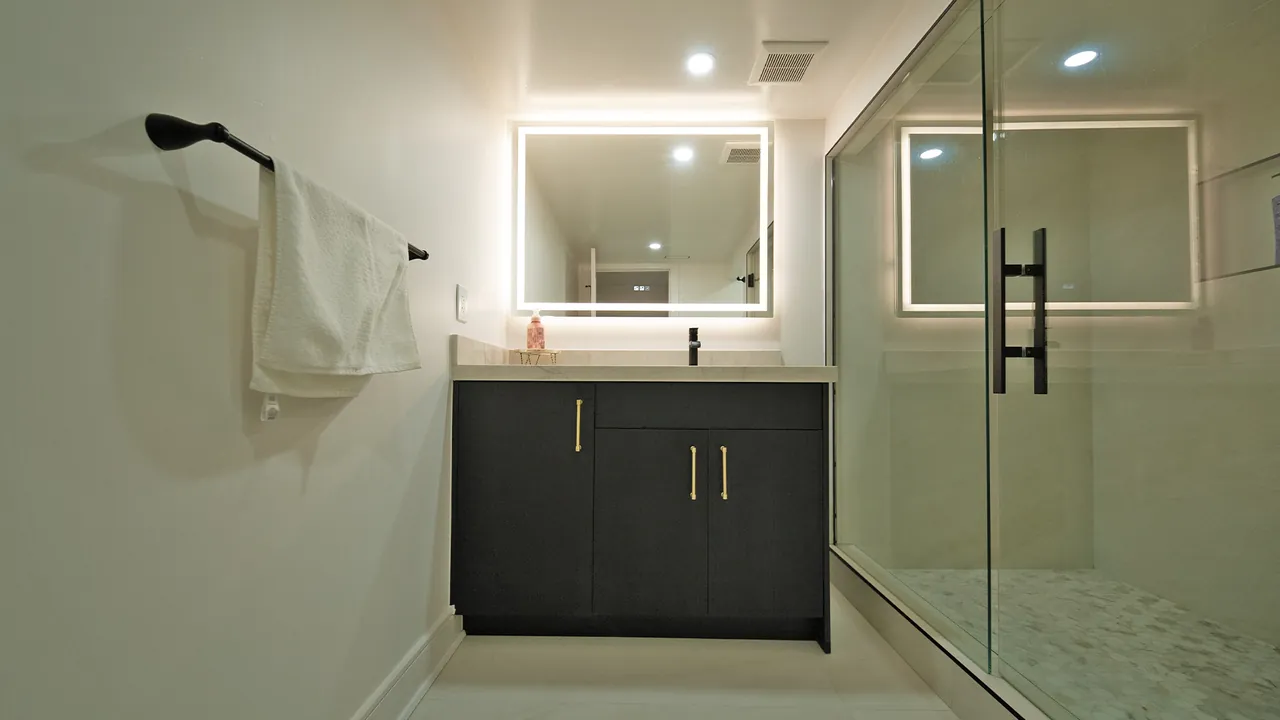Black vanity with a lit mirror beside a glass shower by Mason Oak Carpentry in Detroit, MI